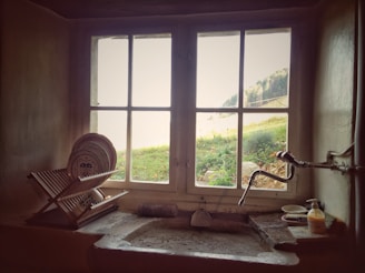 A rustic kitchen scene with a large window providing a view of greenery outside. Inside, an old-fashioned sink area is visible with a dish rack full of plates to the left. The faucet has an antique design, and there are cleaning items neatly placed next to the sink. Natural light softly illuminates the scene, creating a cozy, timeless atmosphere.