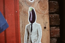 A surreal artwork featuring a collage of a man's body in a suit with an eggplant replacing his head. The background consists of textured wood and brick elements with a red and blue color scheme.