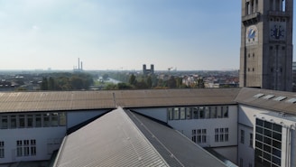 A rooftop view reveals an urban landscape with a prominent tower on the right, displaying large circular dials marked 'Barometer.' In the background, industrial chimneys and a river can be seen, with trees interspersed among the city buildings under a partly cloudy sky.