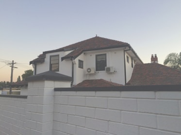A two-story white house with a reddish-brown tiled roof is visible behind a white brick wall. The house features multiple windows framed in black and two air conditioning units mounted on its exterior. In the background, there are utility poles and some trees, while the sky has a gentle gradient from blue to a light orange near the horizon.