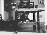A black and white photograph of a child exploring beneath a toy workbench. The child is reaching for a toy tool while surrounded by various toy items. In the background, there are framed photos on the wall and a toy car on the left. The setting appears to be indoors, with wooden flooring and a play-themed environment.