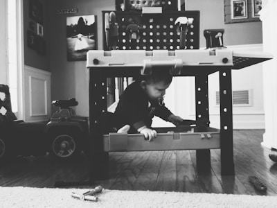 A black and white photograph of a child exploring beneath a toy workbench. The child is reaching for a toy tool while surrounded by various toy items. In the background, there are framed photos on the wall and a toy car on the left. The setting appears to be indoors, with wooden flooring and a play-themed environment.