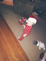 A young child wearing festive red pajamas with white polka dots and a red and white Santa hat stands on a carpeted floor. Nearby are a snowman plush toy and a small gift box with a Santa decoration. The room has a mix of wooden and carpeted flooring.