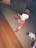 A young child wearing festive red pajamas with white polka dots and a red and white Santa hat stands on a carpeted floor. Nearby are a snowman plush toy and a small gift box with a Santa decoration. The room has a mix of wooden and carpeted flooring.
