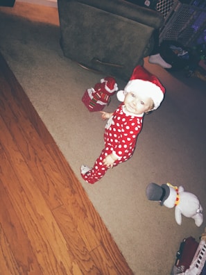 A young child wearing festive red pajamas with white polka dots and a red and white Santa hat stands on a carpeted floor. Nearby are a snowman plush toy and a small gift box with a Santa decoration. The room has a mix of wooden and carpeted flooring.
