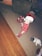 A young child wearing festive red pajamas with white polka dots and a red and white Santa hat stands on a carpeted floor. Nearby are a snowman plush toy and a small gift box with a Santa decoration. The room has a mix of wooden and carpeted flooring.