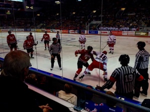 A hockey game is taking place in an indoor arena with players on the ice engaged in an altercation. Two players from opposing teams, one in a red jersey and the other in a white jersey, are involved in a physical fight. There are referees in striped shirts observing the situation closely. In the background, other team members and spectators are visible, creating an energetic atmosphere.