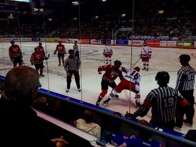 A hockey game is taking place in an indoor arena with players on the ice engaged in an altercation. Two players from opposing teams, one in a red jersey and the other in a white jersey, are involved in a physical fight. There are referees in striped shirts observing the situation closely. In the background, other team members and spectators are visible, creating an energetic atmosphere.