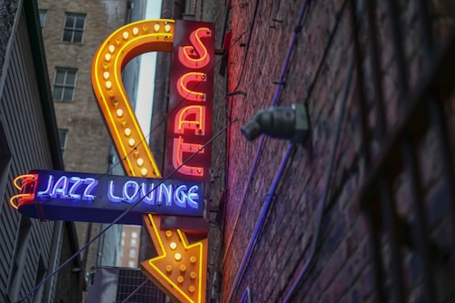 A brightly lit neon sign with the words 'JAZZ LOUNGE' in blue and 'SCAT' in red, outlined by a large yellow arrow. The sign is mounted on a brick wall in a narrow alleyway, and the perspective angles upwards, showing part of the building and some exposed pipes.