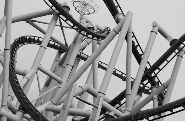 A complex and intricate structure composed of numerous interwoven steel beams and roller coaster tracks. The image is in black and white, emphasizing the geometric patterns and industrial design. The tracks loop and twist, creating a sense of motion and engineering marvel.