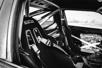 The interior of a stripped-down car, possibly a racing vehicle, featuring two bucket seats with harnesses. The seats are labeled with 'Sparco' and 'Bride' branding. The car lacks typical interior finishes, exposing the metal framework and roll cage.