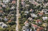 An aerial view of a suburban neighborhood with numerous houses featuring red and gray rooftops. The area is lush with greenery, as trees and gardens are visible around the properties. A main road runs through the middle of the image, lined with houses on both sides, and several cars are parked along the road.