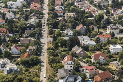 An aerial view of a suburban neighborhood with numerous houses featuring red and gray rooftops. The area is lush with greenery, as trees and gardens are visible around the properties. A main road runs through the middle of the image, lined with houses on both sides, and several cars are parked along the road.