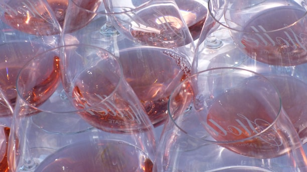Several wine glasses are filled with a pinkish rosé wine, with reflections and refractions of light creating a shimmering effect. The glasses appear stacked or clustered together, showcasing an elegant arrangement with visible branding on the glass.