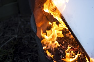 Bright orange flames are dancing around charred wood and material inside a barrel or container. The fire creates an intense and chaotic energy, with embers and ashes scattered around. A hard, light-colored surface is partially visible reflecting the fiery glow.