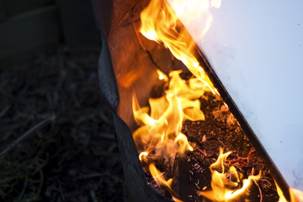 Bright orange flames are dancing around charred wood and material inside a barrel or container. The fire creates an intense and chaotic energy, with embers and ashes scattered around. A hard, light-colored surface is partially visible reflecting the fiery glow.