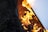 Bright orange flames are dancing around charred wood and material inside a barrel or container. The fire creates an intense and chaotic energy, with embers and ashes scattered around. A hard, light-colored surface is partially visible reflecting the fiery glow.