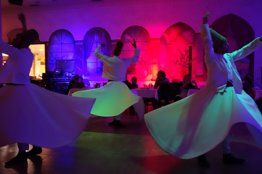 A group of Sufi whirling dervishes in motion, with vibrant, colorful lighting casting hues of purple, pink, and blue across the scene. The performers wear traditional white garments and tall hats, creating a mesmerizing, circular motion that evokes spirituality and trance. In the background, seated people observe the performance in a dimly lit, arched venue.