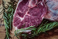 A close-up view of a raw beef steak placed on a wooden surface, surrounded by sprigs of fresh rosemary. The steak is marbled and has visible bone and fat, indicating a tender cut of meat.