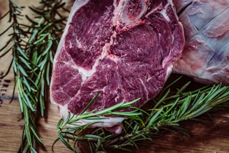 A close-up view of a raw beef steak placed on a wooden surface, surrounded by sprigs of fresh rosemary. The steak is marbled and has visible bone and fat, indicating a tender cut of meat.