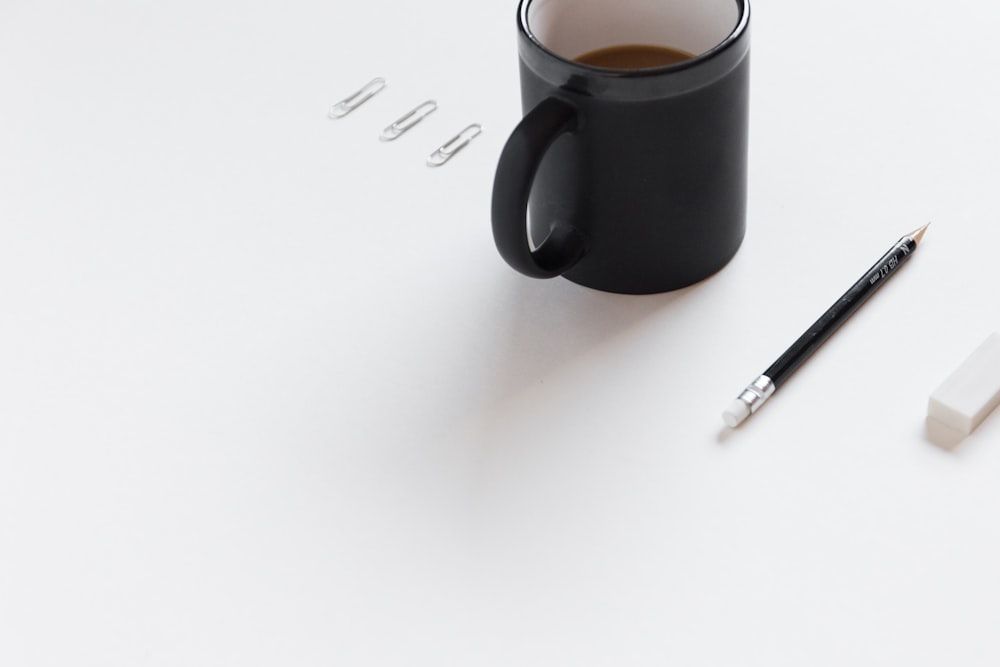 trois trombones blancs, une tasse en céramique noire, un crayon noir et une gomme blanche sur une surface blanche