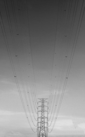 A high voltage transmission tower is captured from an upward angle, with numerous power lines extending towards the sky, creating a converging perspective. The image is in black and white, emphasizing the linear patterns of the cables against a cloudy sky.