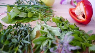 Various fresh herbs and a halved red bell pepper are arranged on a wooden surface. The herbs include mint, chives, and parsley, showcasing a vibrant mix of green hues.
