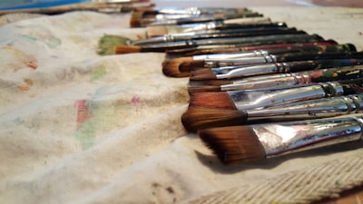 A collection of paintbrushes lies neatly arranged on a paint-splattered cloth. The brushes have different colored bristles and paint-stained handles, reflecting signs of frequent use. The scene suggests an artist's workspace.