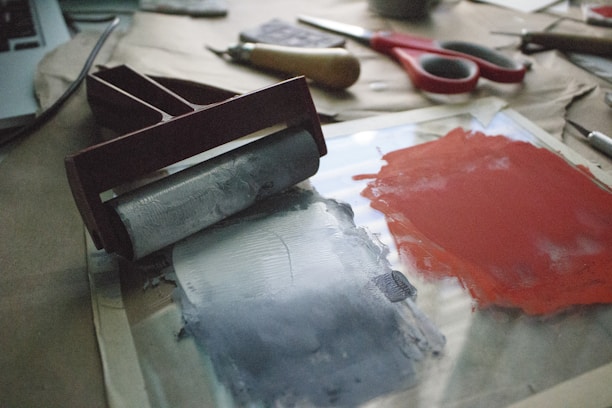 A workspace featuring printing tools with a brayer resting on a glass surface next to patches of red and gray ink. The background includes a pair of scissors, additional tools, and a laptop, suggesting a creative process in progress.