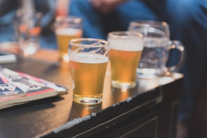 Several glasses of beer are placed on a dark coffee table with magazines. The background is slightly blurred, showing what appears to be a social setting.