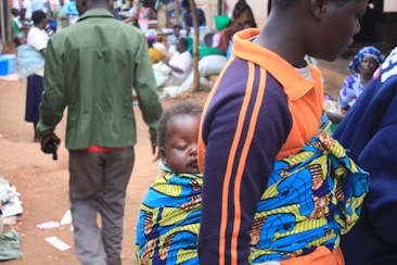 A young child is securely wrapped on the back of a person wearing a brightly colored print fabric. The child is asleep and appears comfortable. The scene is set in what seems to be a busy outdoor market or public area with several people in the background. Some individuals are engaged in different activities, and there are scattered items on the ground.