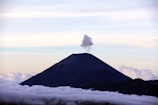 A majestic volcano rises above a thick layer of clouds, with a plume of smoke or vapor emitting from its peak against a serene sky.