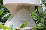 A large industrial or mechanical structure with a cylindrical base is surrounded by lush green foliage. The structure has a central round element with several bolts and is connected by black cables. Large tropical leaves frame the scene, indicating a blend of industrial and natural elements.