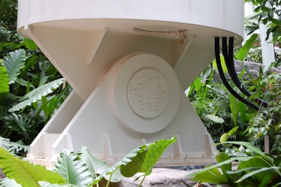 A large industrial or mechanical structure with a cylindrical base is surrounded by lush green foliage. The structure has a central round element with several bolts and is connected by black cables. Large tropical leaves frame the scene, indicating a blend of industrial and natural elements.