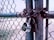 A chain-link fence is securely fastened with a heavy metal chain and two padlocks. The metal of the fence and locks shows signs of rust and weathering. The background depicts an open outdoor space with patches of grass and dirt.