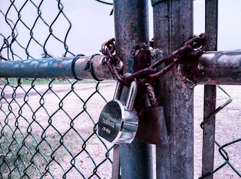 A chain-link fence is securely fastened with a heavy metal chain and two padlocks. The metal of the fence and locks shows signs of rust and weathering. The background depicts an open outdoor space with patches of grass and dirt.