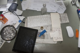 A cluttered desk features a variety of items including a keyboard, mouse, tablet, notebook, and several papers scattered around. There's a noticeable ashtray with cigarette butts, a lighter, a bottle, and pens. The surface is covered with documents, some crumpled, and various office supplies.