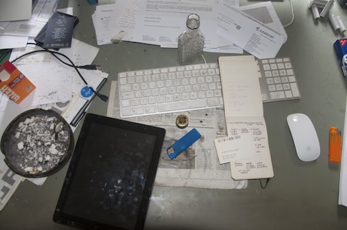 A cluttered desk features a variety of items including a keyboard, mouse, tablet, notebook, and several papers scattered around. There's a noticeable ashtray with cigarette butts, a lighter, a bottle, and pens. The surface is covered with documents, some crumpled, and various office supplies.