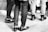 A black and white image capturing the legs and feet of a group of people standing together. They are wearing various types of footwear ranging from casual to formal, and the ground is covered with brick tiles.