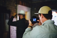Two individuals are in a dimly lit room. One person is holding a camera, capturing a photo of the other who is adjusting their hat. The setting has a rustic feel with brick walls and a partially lit window.