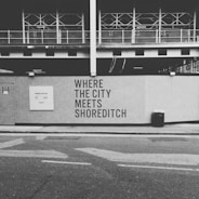 A black and white image of a construction site with a wall featuring the text 'WHERE THE CITY MEETS SHOREDITCH' prominently displayed. Above the wall, there is a partially constructed building with metal framework visible. The street in the foreground includes a trash bin and a sign with website information.