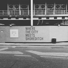 A black and white image of a construction site with a wall featuring the text 'WHERE THE CITY MEETS SHOREDITCH' prominently displayed. Above the wall, there is a partially constructed building with metal framework visible. The street in the foreground includes a trash bin and a sign with website information.