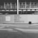A black and white image of a construction site with a wall featuring the text 'WHERE THE CITY MEETS SHOREDITCH' prominently displayed. Above the wall, there is a partially constructed building with metal framework visible. The street in the foreground includes a trash bin and a sign with website information.