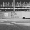 A black and white image of a construction site with a wall featuring the text 'WHERE THE CITY MEETS SHOREDITCH' prominently displayed. Above the wall, there is a partially constructed building with metal framework visible. The street in the foreground includes a trash bin and a sign with website information.