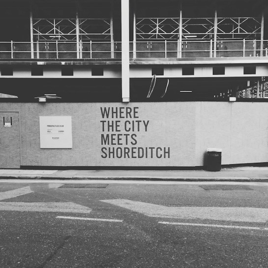 A black and white image of a construction site with a wall featuring the text 'WHERE THE CITY MEETS SHOREDITCH' prominently displayed. Above the wall, there is a partially constructed building with metal framework visible. The street in the foreground includes a trash bin and a sign with website information.