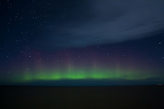 A nighttime sky displaying an aurora with greenish hues near the horizon that blend into a dark sky filled with stars. A large expanse of clouds is visible occupying the upper sky.