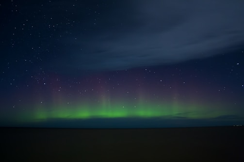 A nighttime sky displaying an aurora with greenish hues near the horizon that blend into a dark sky filled with stars. A large expanse of clouds is visible occupying the upper sky.