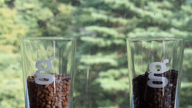 Two clear glass containers are filled with coffee beans. The glass on the left contains lighter brown beans, while the glass on the right has darker roasted beans. Each glass has a white 'g' logo on it. The background shows an out-of-focus view of green foliage, suggesting a natural setting.