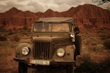 An old military-style jeep is parked on a grassy, rocky terrain with dramatic, rugged, brownish-red rock formations in the background under a cloudy sky.