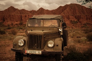 An old military-style jeep is parked on a grassy, rocky terrain with dramatic, rugged, brownish-red rock formations in the background under a cloudy sky.